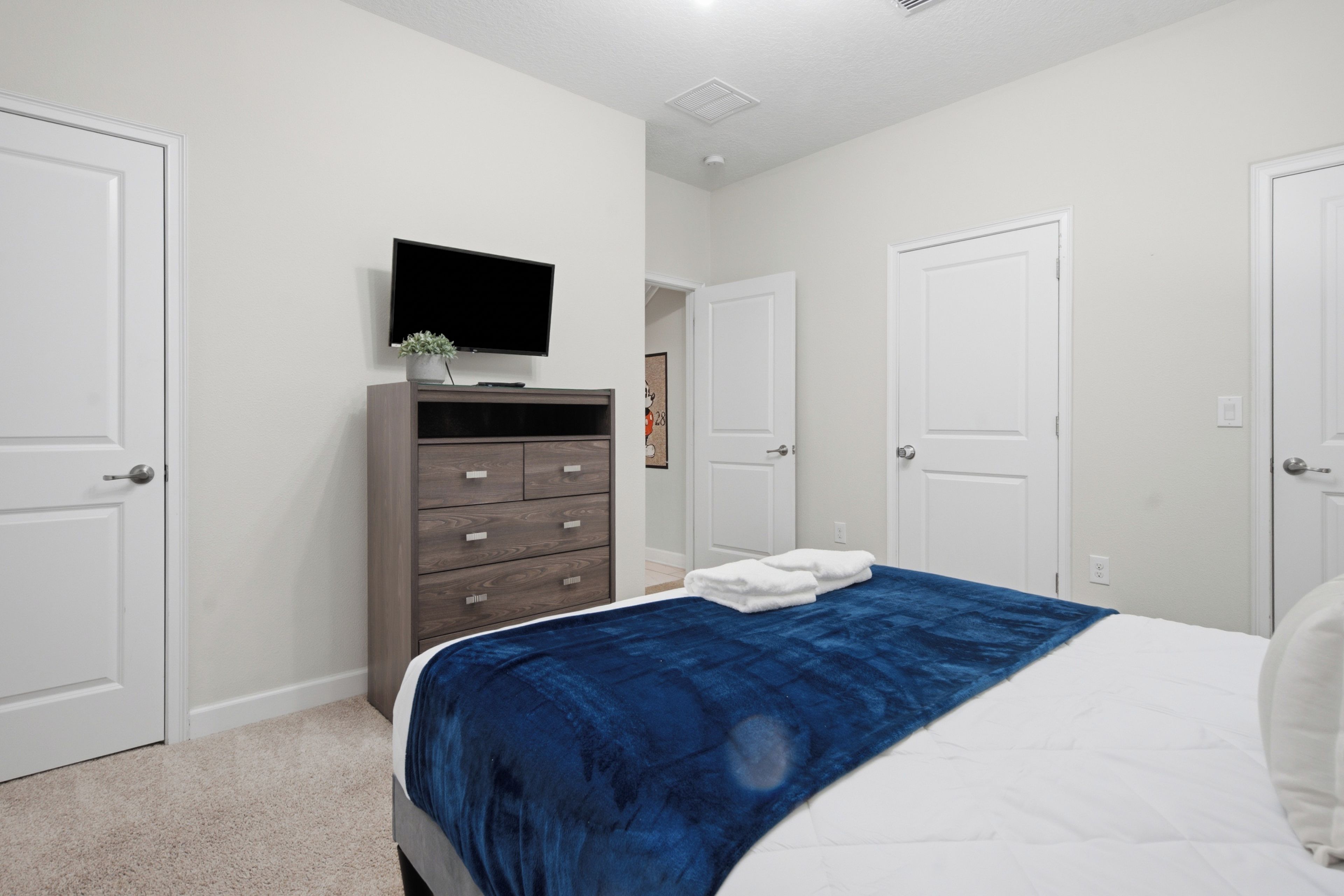 Une chambre à coucher avec un lit, une commode, un téléviseur fixé au mur et deux portes.