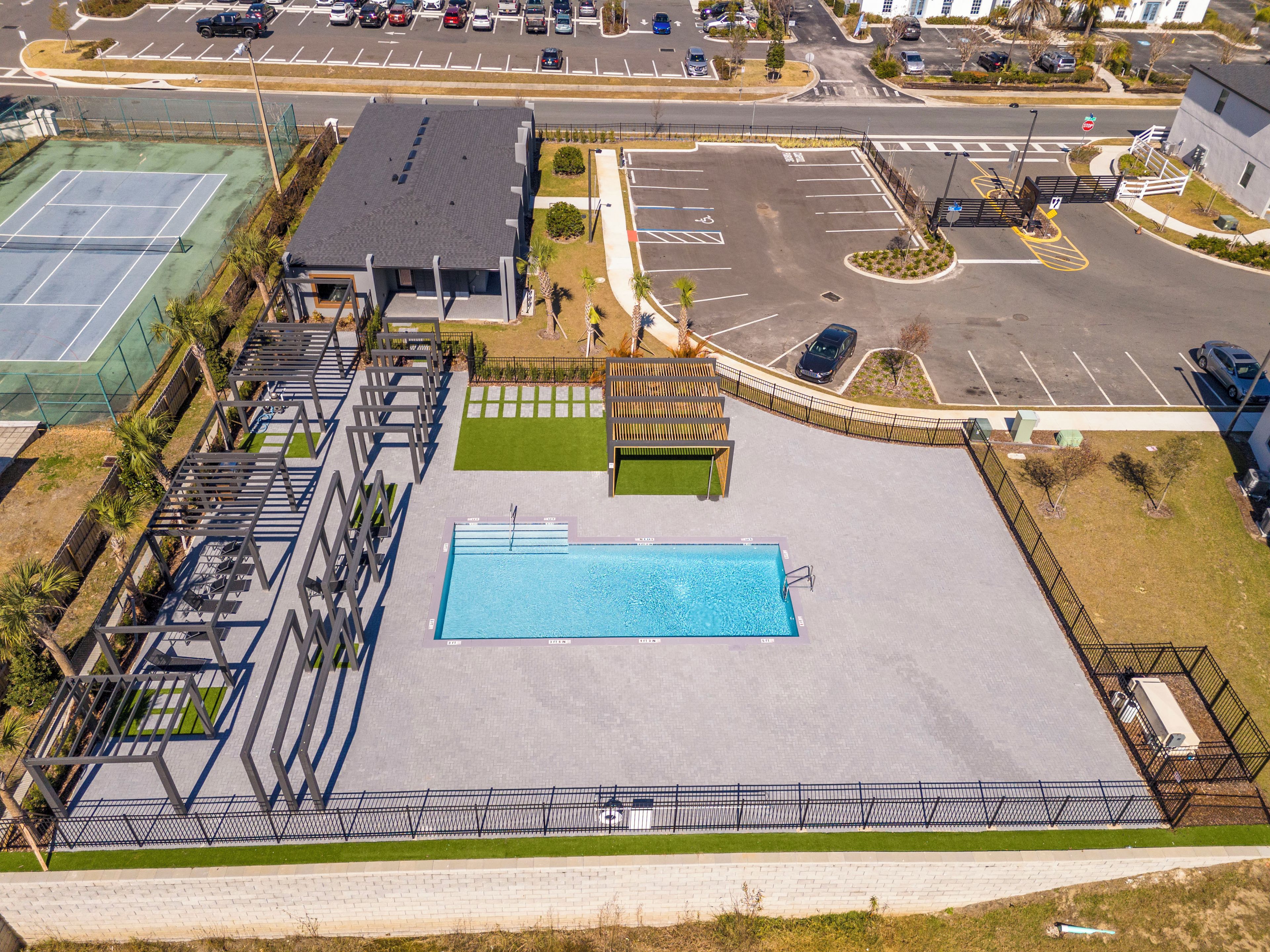 Ein Schwimmbad mit umlaufender Terrasse, ein Parkplatz und ein Gebäude mit flachem Dach.