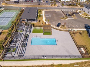 Ein Schwimmbad mit umlaufender Terrasse, ein Parkplatz und ein Gebäude mit flachem Dach.
