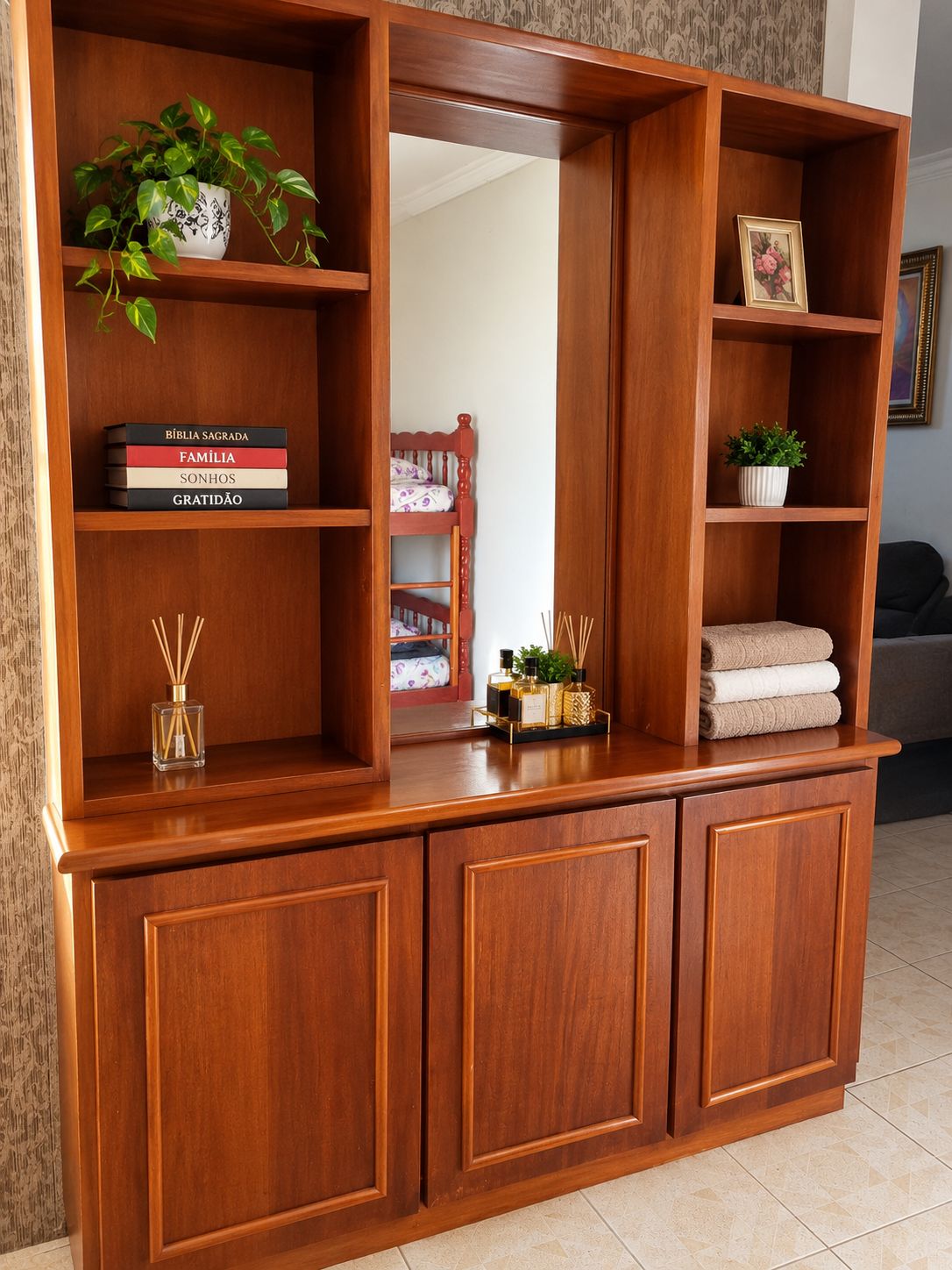 Um console de madeira com prateleiras contendo livros, uma planta, um quadro emoldurado e uma pequena mesa com um frasco e difusor de vime.