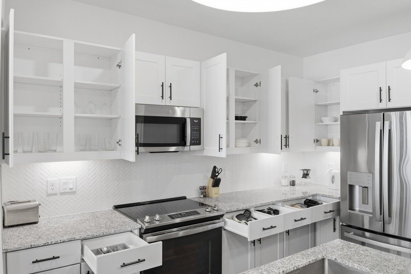 A modern kitchen with white cabinetry, a built-in microwave, a stainless steel refrigerator, and a granite countertop.
