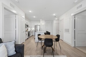 A modern kitchen with white cabinetry, a central island, and a dining area with a round table and chairs. - Beautiful 2BR near PGA and Universal Studios (Frisco)