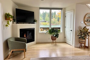 Ein Wohnzimmer mit Kamin, Fernseher und einem großen Fenster mit Blick auf Bäume und einen Steg.
