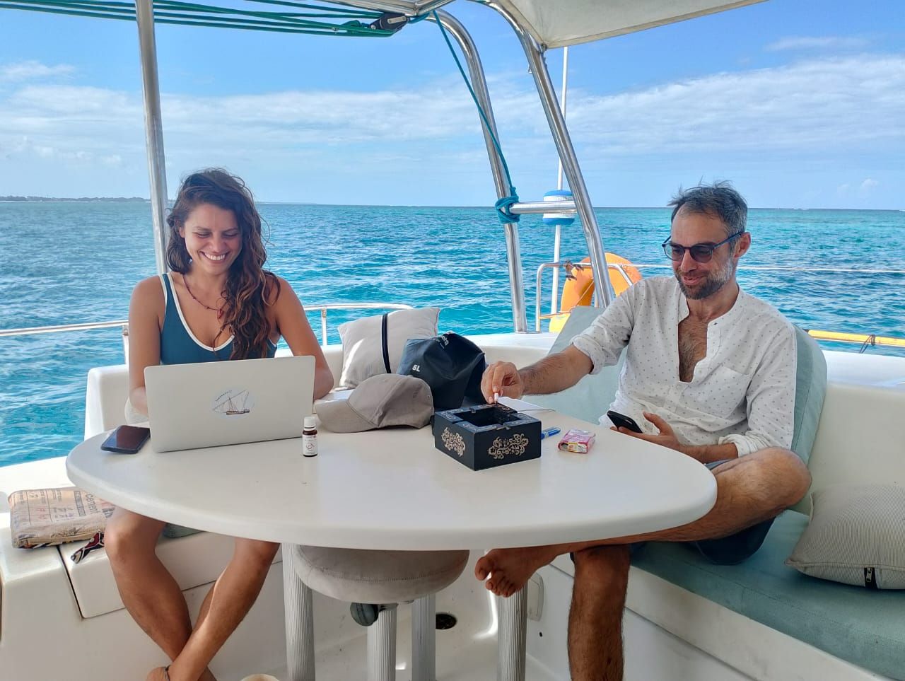 Dos personas sentadas a una mesa redonda en un barco, con una computadora portátil y una caja de pañuelos.