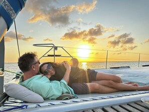 Una pareja descansando en una tumbona en la cubierta de un barco, con un atardecer de fondo.
