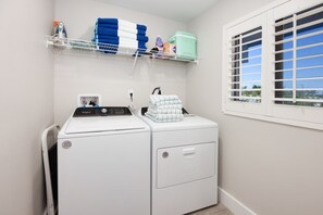 A laundry area with a front-loading washer and dryer, a window with blinds, and a shelf with folded towels and laundry supplies. - Pelicans Roost 303 – Beautifully Remodeled Beachfront Escape with Gulf Views & H (Sanibel)