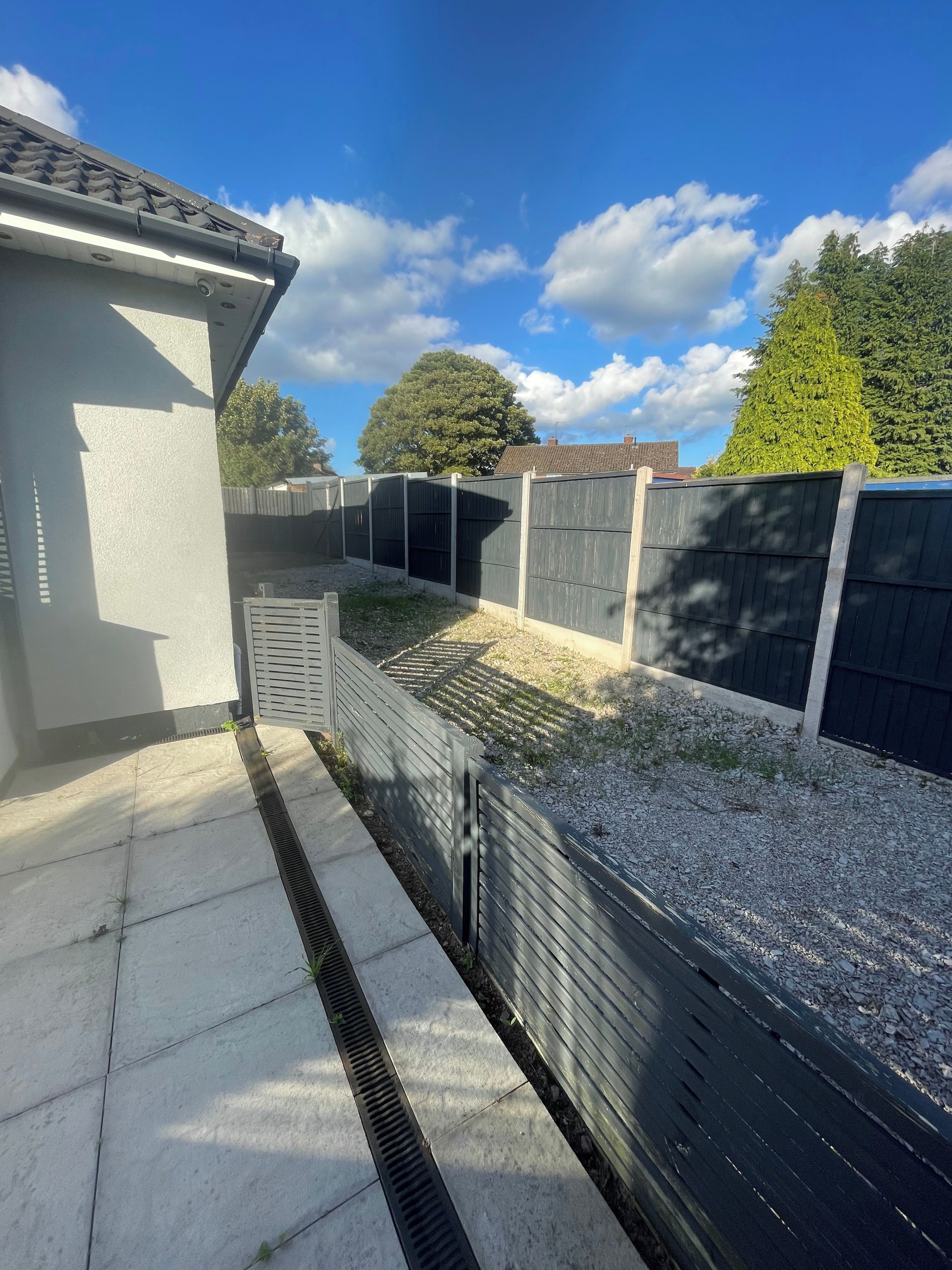 A residential backyard with a paved area, a metal fence, and a tree in the background.