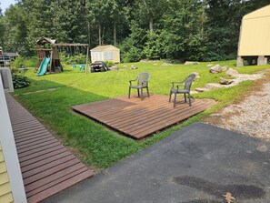 Ein Garten mit einer Holzterrasse, zwei Stühlen, einem Spielbereich mit einer Rutsche und einem Geräteschuppen.