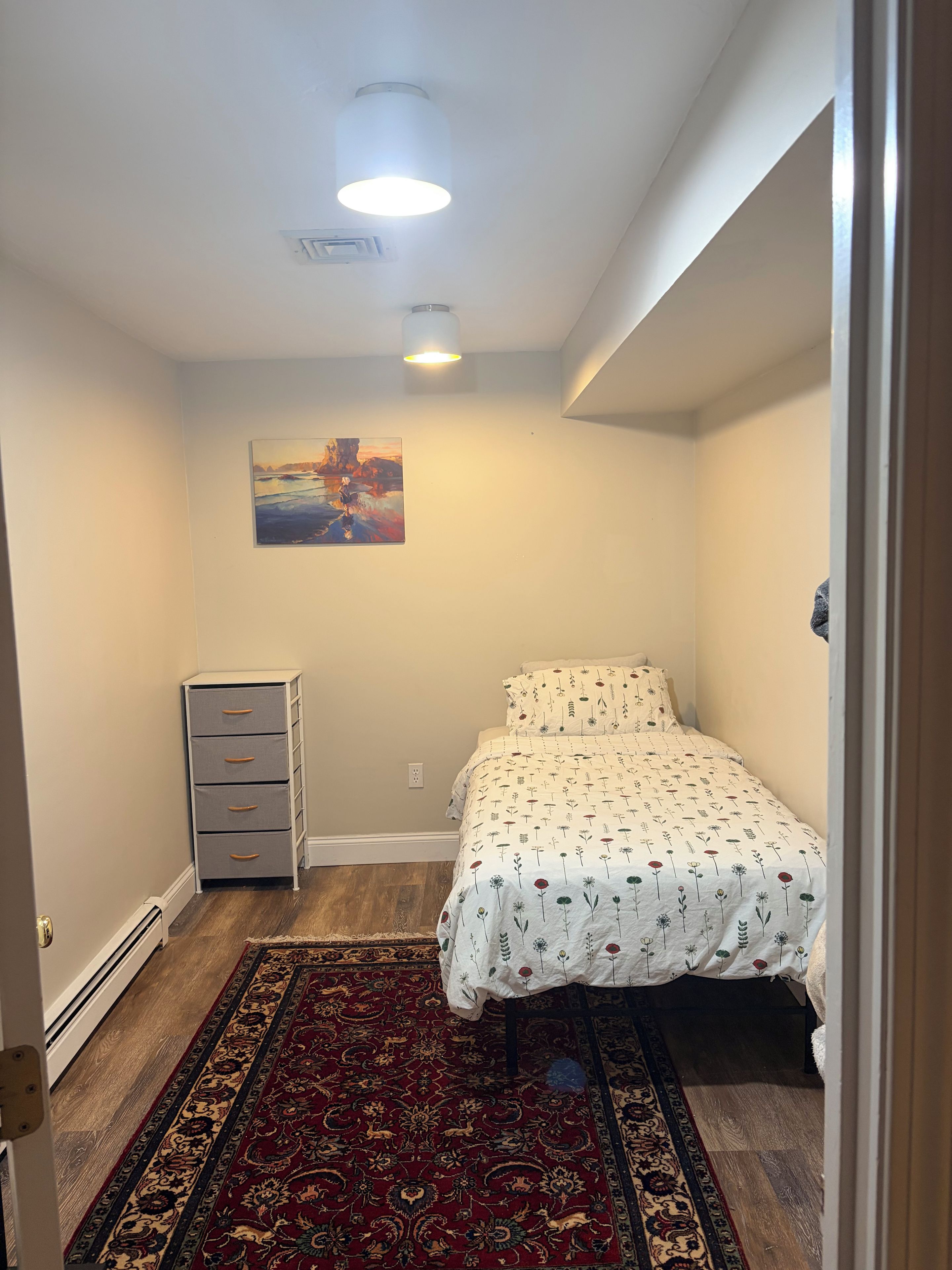 Une chambre à coucher avec un lit, une table de chevet, une commode, un tapis et un tableau accroché au mur.