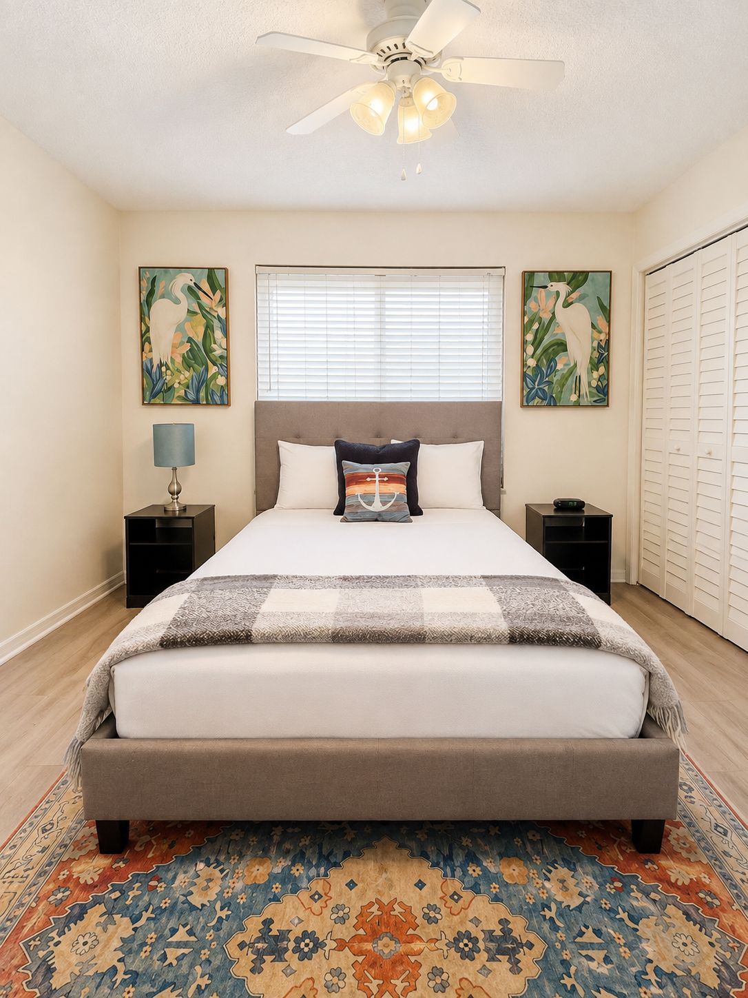 A bedroom with a bed, a ceiling fan, two nightstands, a lamp, a window with blinds, and two wall-mounted paintings.