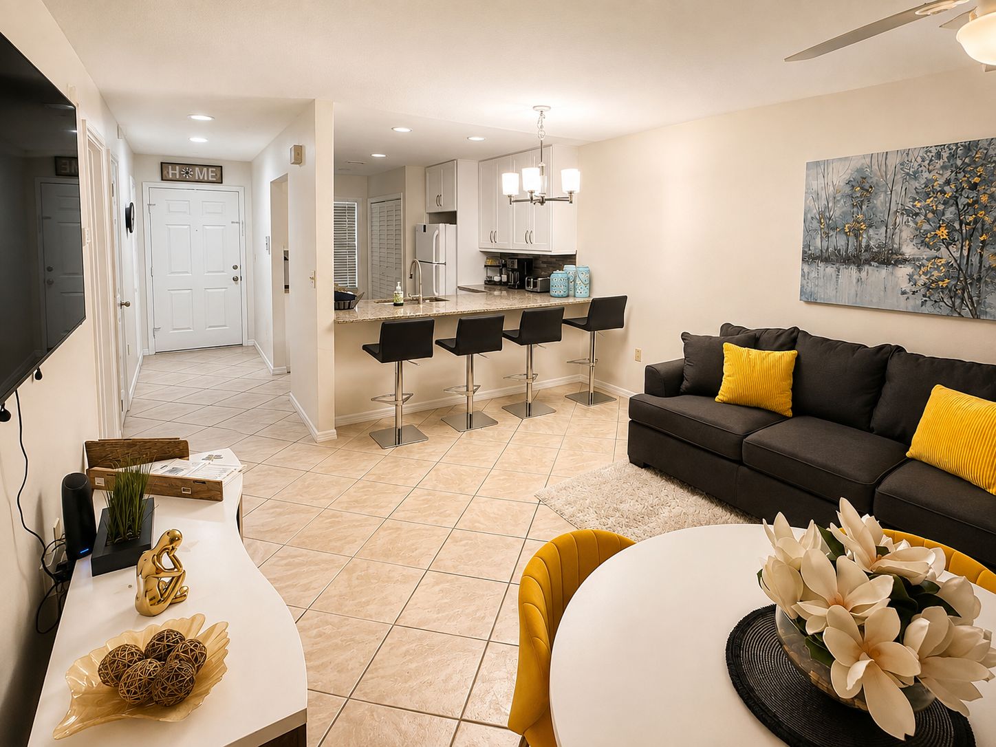 A modern living room with a sofa, a dining area with chairs, a kitchen with bar stools, and a hallway leading to a door marked 'HOME'.