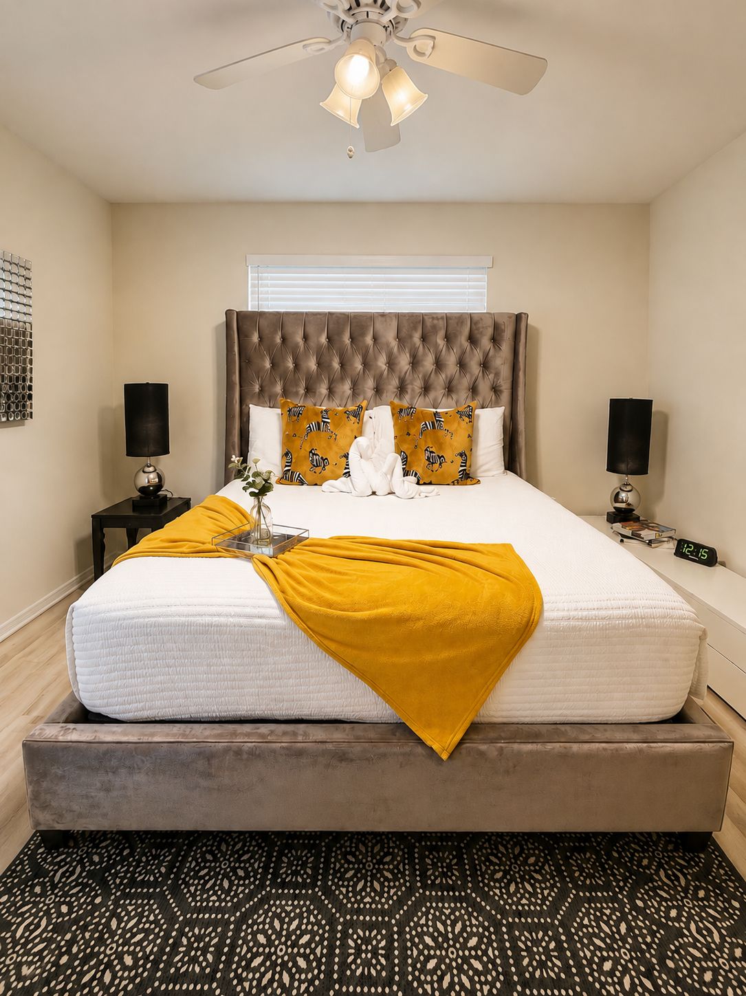 Un dormitorio con cabecera capitonada, una cama con colchón blanco y una manta amarilla, dos mesitas de noche con lámparas, un reloj y una alfombra estampada.