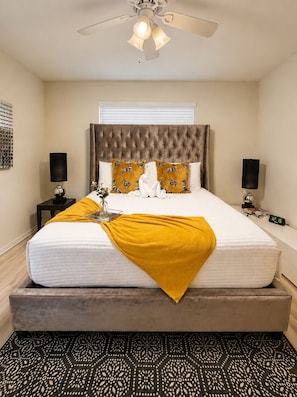 A bedroom with a tufted headboard, a bed with a white mattress and a yellow throw, two bedside tables with lamps, a clock, and a patterned rug.