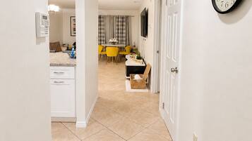 A hallway with a kitchen area, a dining table, and a clock on the wall.