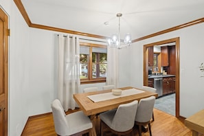 A dining area with a wooden table and chairs, a chandelier, and a view of the kitchen through an open door.