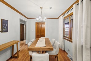 A dining room with a wooden table, white chairs, a chandelier, and a window with curtains.