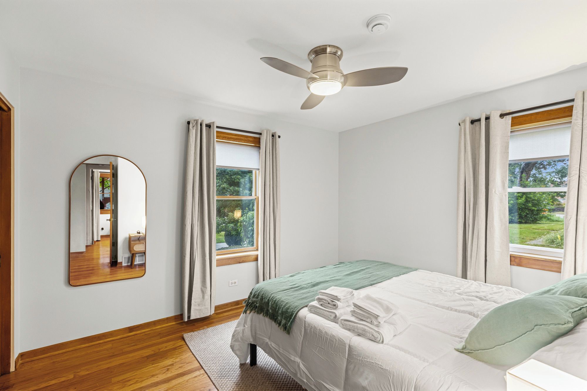 Une chambre à coucher avec un lit, un ventilateur de plafond, un miroir et deux fenêtres avec des rideaux.
