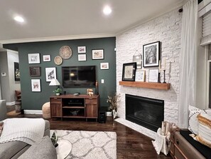 A living room with a fireplace, a TV mounted on the wall, and a variety of framed pictures. - Luxurious Retreat: 3-Bed/3-Bath Condo (Chicago)