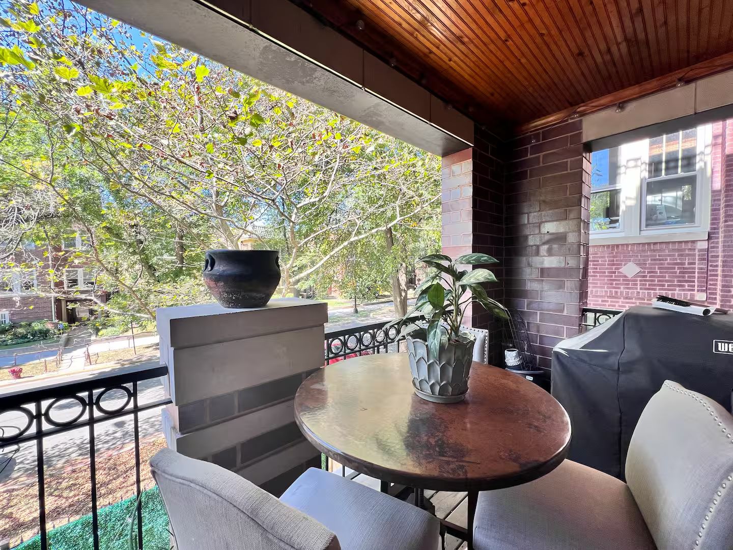 An outdoor seating area with a table and chairs, a potted plant, and a view of a street and trees.
