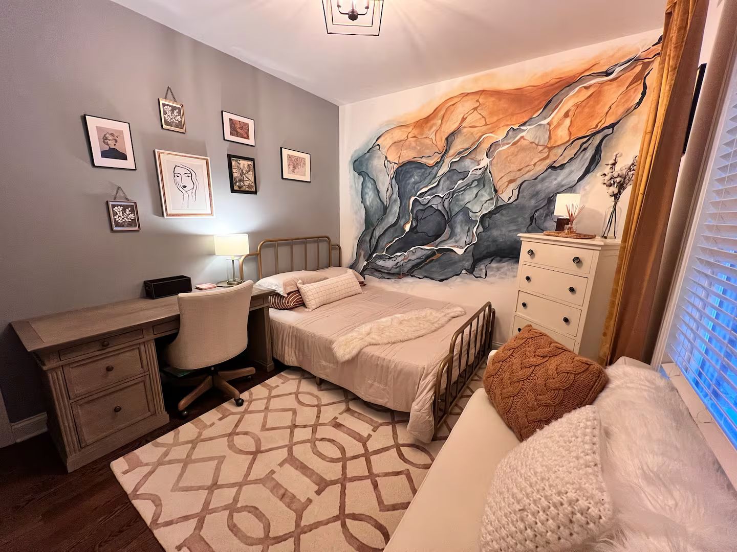 A bedroom with a bed, desk, chair, dresser, and a large abstract painting on the wall.