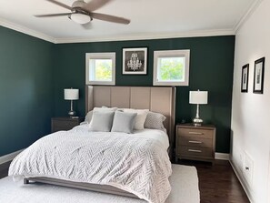 Ein Schlafzimmer mit einem Bett, zwei Nachttischen, einem Deckenventilator und zwei mit Vorhängen versehenen Fenstern.