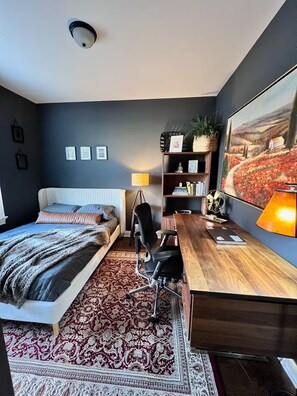 Ein Schlafzimmer mit Bett, Schreibtisch, Stuhl, Bücherregal und einem großen Gemälde an der Wand.