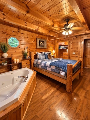 A wooden cabin bedroom with a large bed, a wooden floor, a ceiling fan, and a window with a view of trees. - A Piece of Paradise| Pigeon Forge Cabin| Hot Tub (Sevier County)