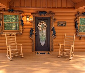 L’entrée d’un chalet en bois, avec une porte sombre, deux fauteuils à bascule, une pancarte de bienvenue et une fenêtre donnant sur les arbres.