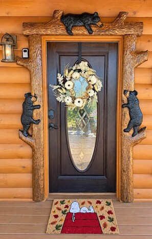 Une porte en bois menant à un chalet, ornée de sculptures d’ours de chaque côté, avec une guirlande au-dessus de la porte et un paillasson décoré d’un motif de Snoopy.
