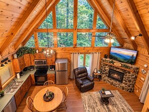 A cozy cabin interior with a kitchen, dining area, and living room, featuring wooden walls, a stone fireplace, and a large window overlooking a forest. - A Piece of Paradise| Pigeon Forge Cabin| Hot Tub (Sevier County)