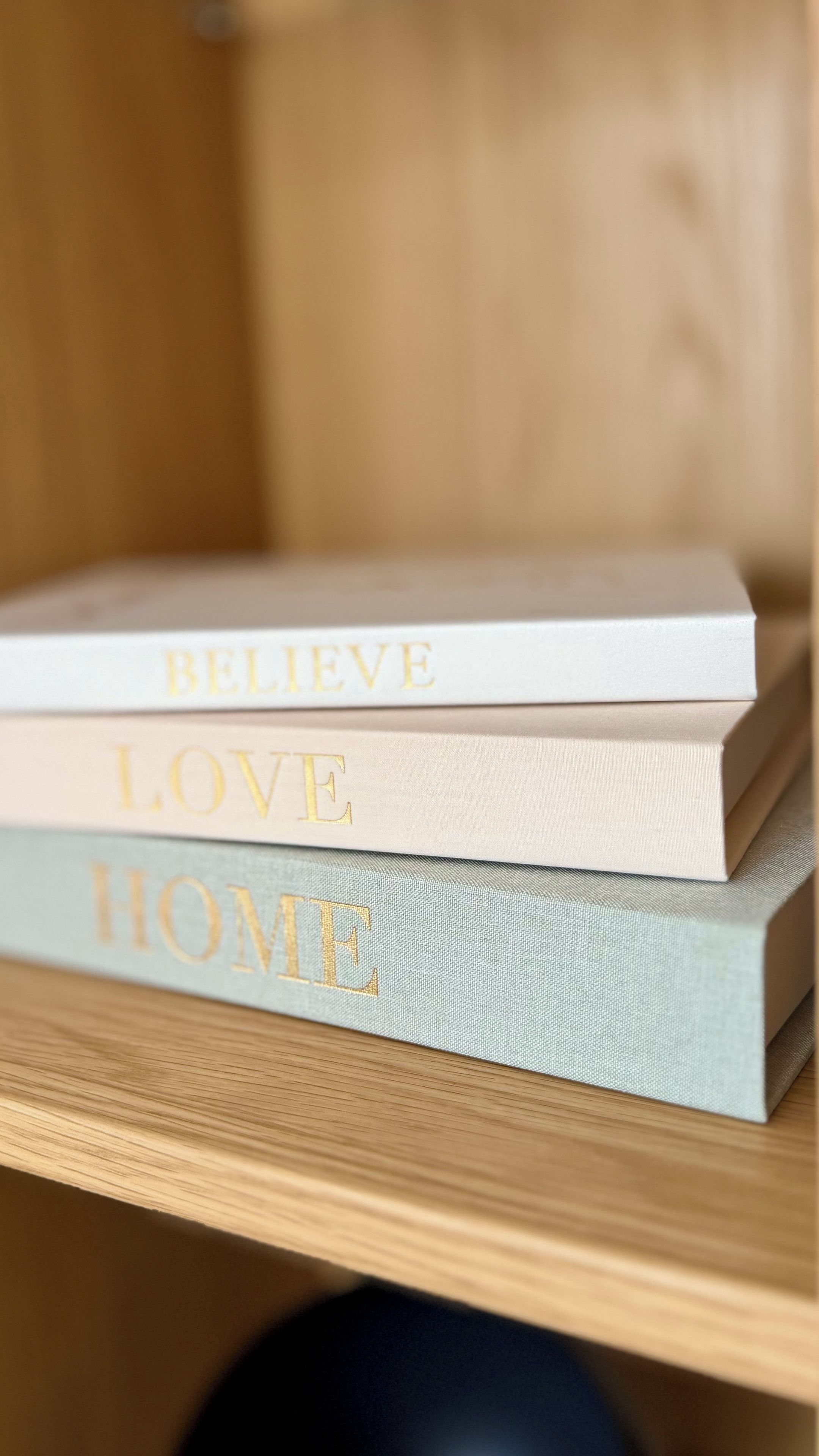 Eine Reihe Bücher mit den Titeln 'BELIEVE', 'LOVE' und 'HOME' auf einem Holzregal.