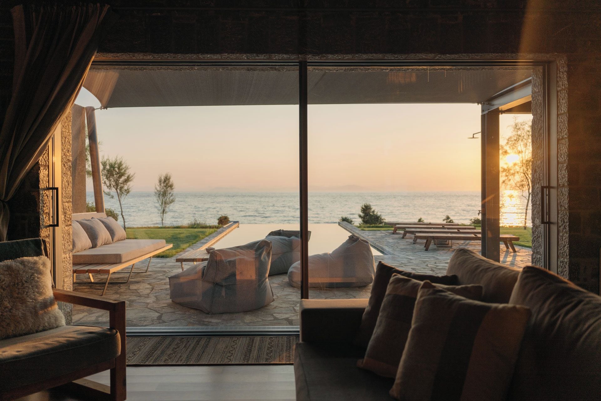 A room with a large glass door leading to a patio with outdoor seating and a view of the sea.