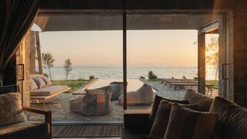 A room with a large glass door leading to a patio with outdoor seating and a view of the sea.