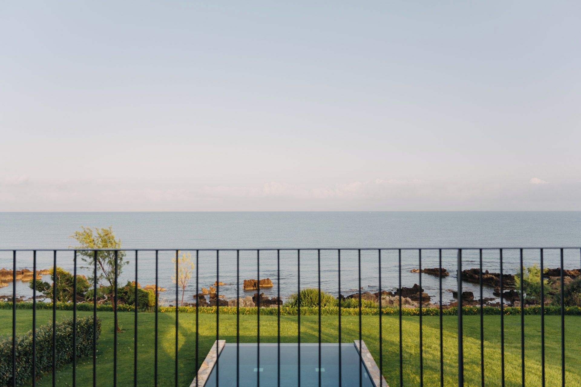 Piscina con vista al mar, rodeada de césped y una valla metálica.