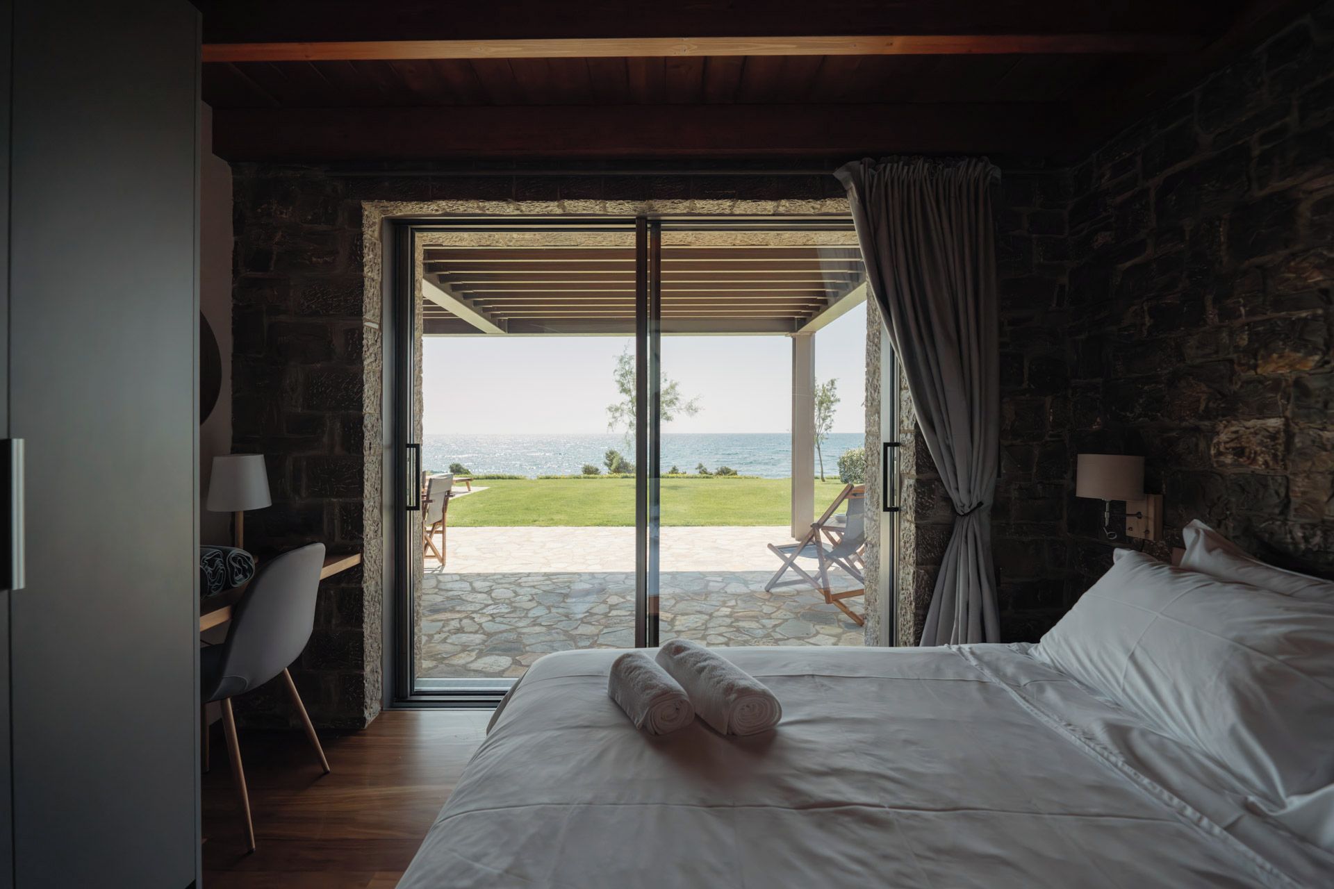 A bedroom with a bed, a chair, a table, and a view of the sea through an open door.
