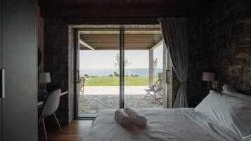 A bedroom with a bed, a chair, a table, and a view of the sea through an open door.