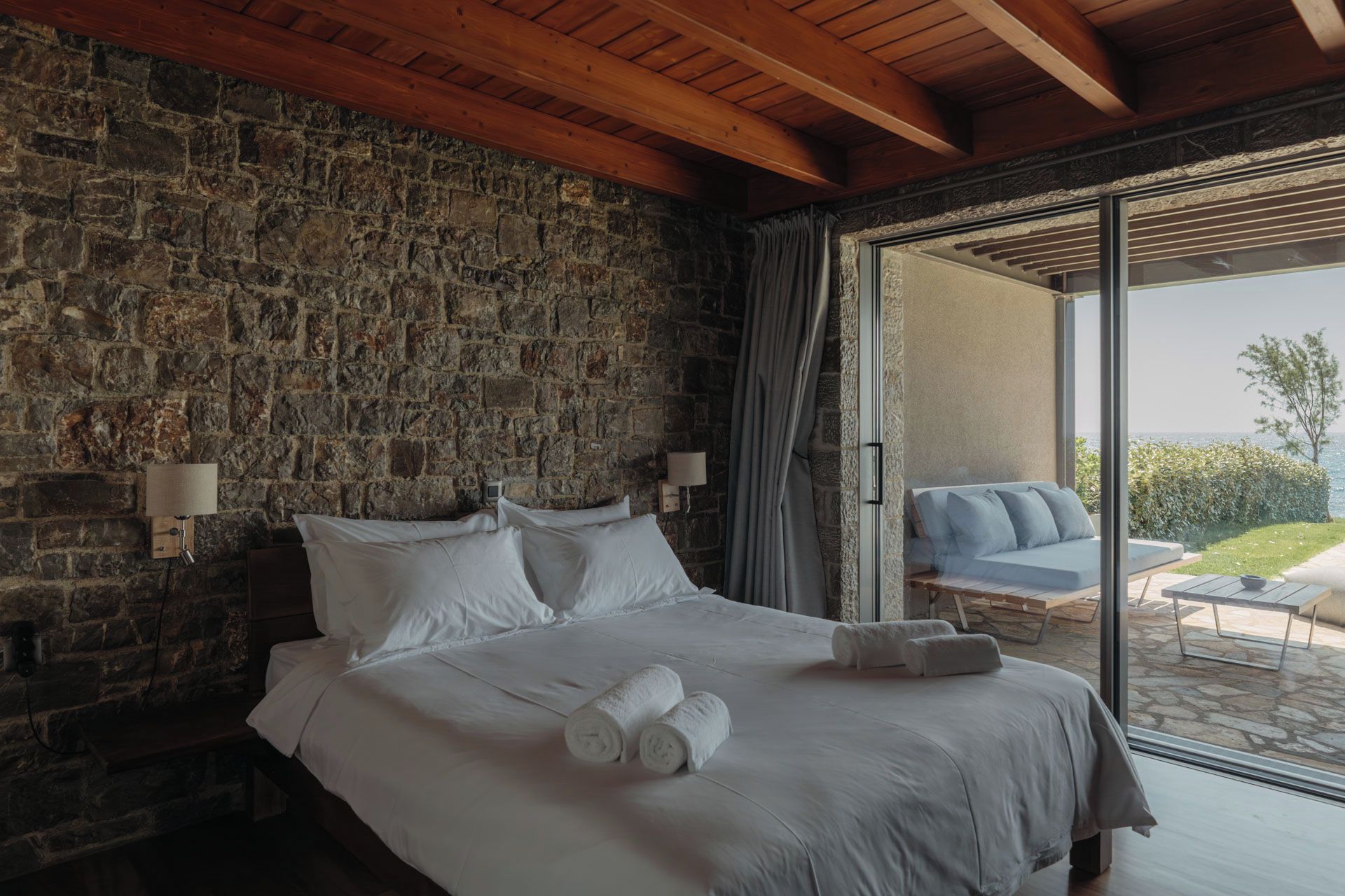 A bedroom with a stone wall, a wooden ceiling, a bed with white linens, a nightstand, a lamp, and a view of a patio with a sofa.