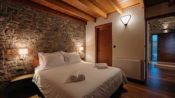 Un dormitorio con pared de piedra, vigas de madera en el techo, una cama con sábanas blancas, dos lámparas de noche y una puerta de madera.