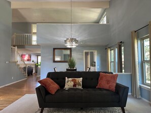 A spacious living room with a black sofa, two decorative pillows, a hanging chandelier, and a view of a kitchen and staircase.