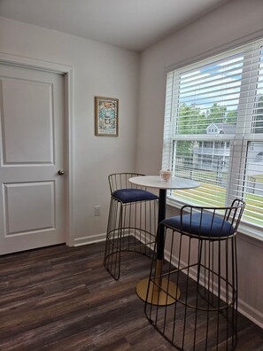 A room with a door, a framed picture on the wall, a window with blinds, a round table with a chair, and a view of a house through the window. - NEW corner lot FURNISHED 3 BR, 2.5 bath townhouse N Raleigh w/security/Internet. (Raleigh)