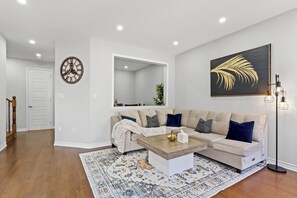 A living room with a sectional sofa, a coffee table, a patterned rug, a wall clock, a framed artwork, and recessed ceiling lights.