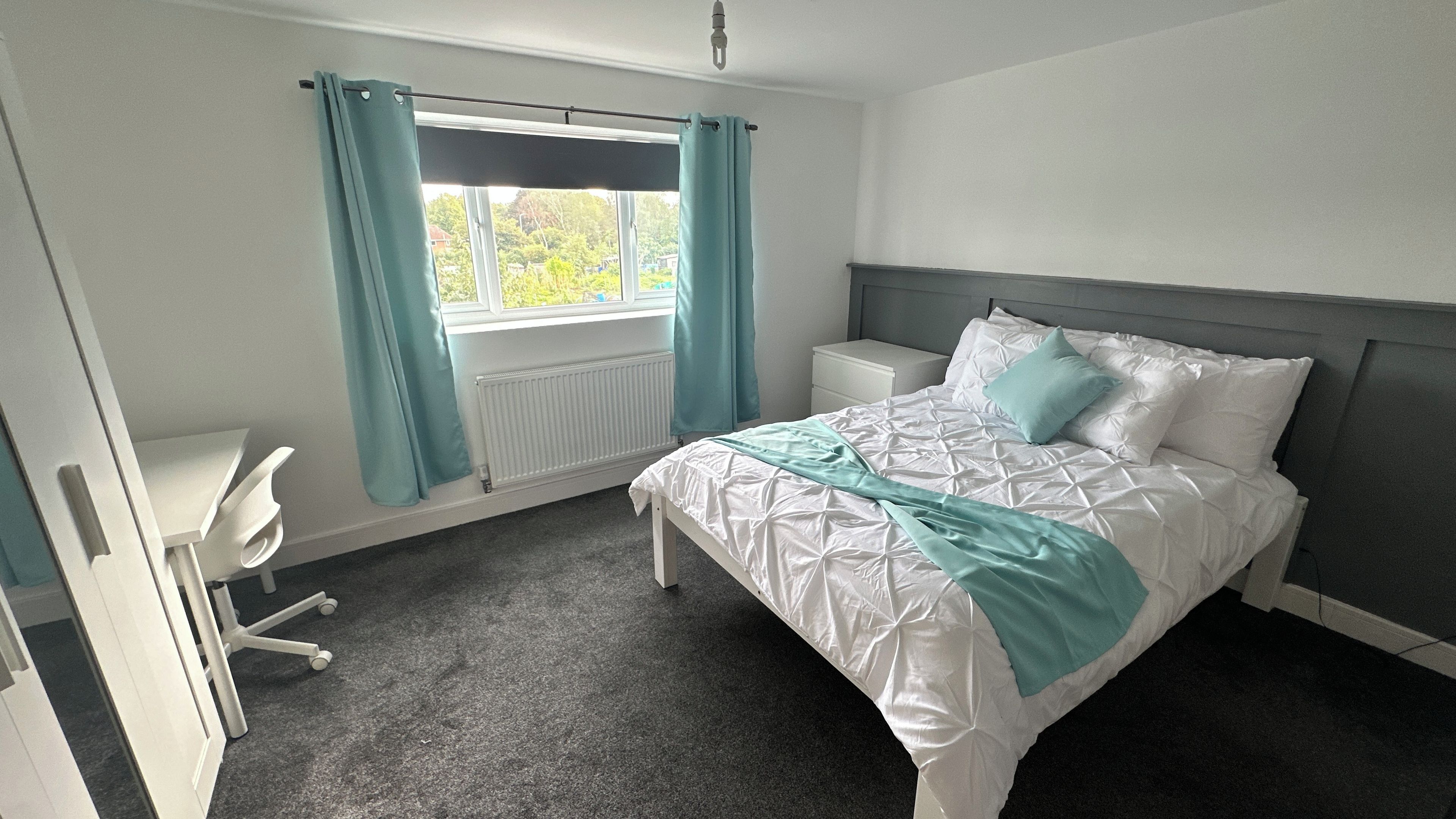 A bedroom with a bed, a desk, a chair, a window with curtains, and a radiator.