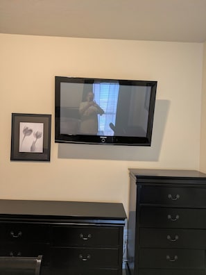 Ein Zimmer mit einem wandmontierten Flachbildfernseher, einem gerahmten Bild an der Wand und einer schwarzen Kommode mit Schubladen.