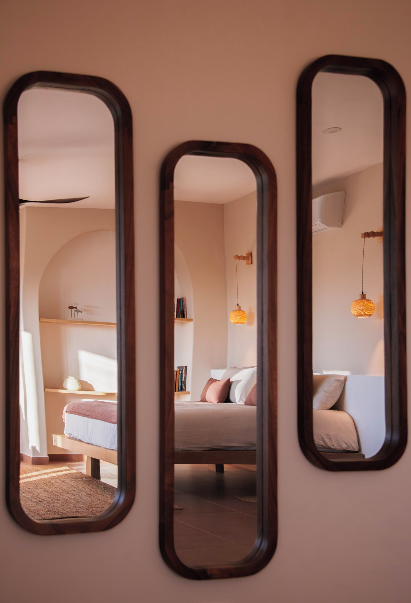 Drei Spiegel an einer Wand, die ein Schlafzimmer mit Bett, Kissen und einer Hängelampe widerspiegeln.