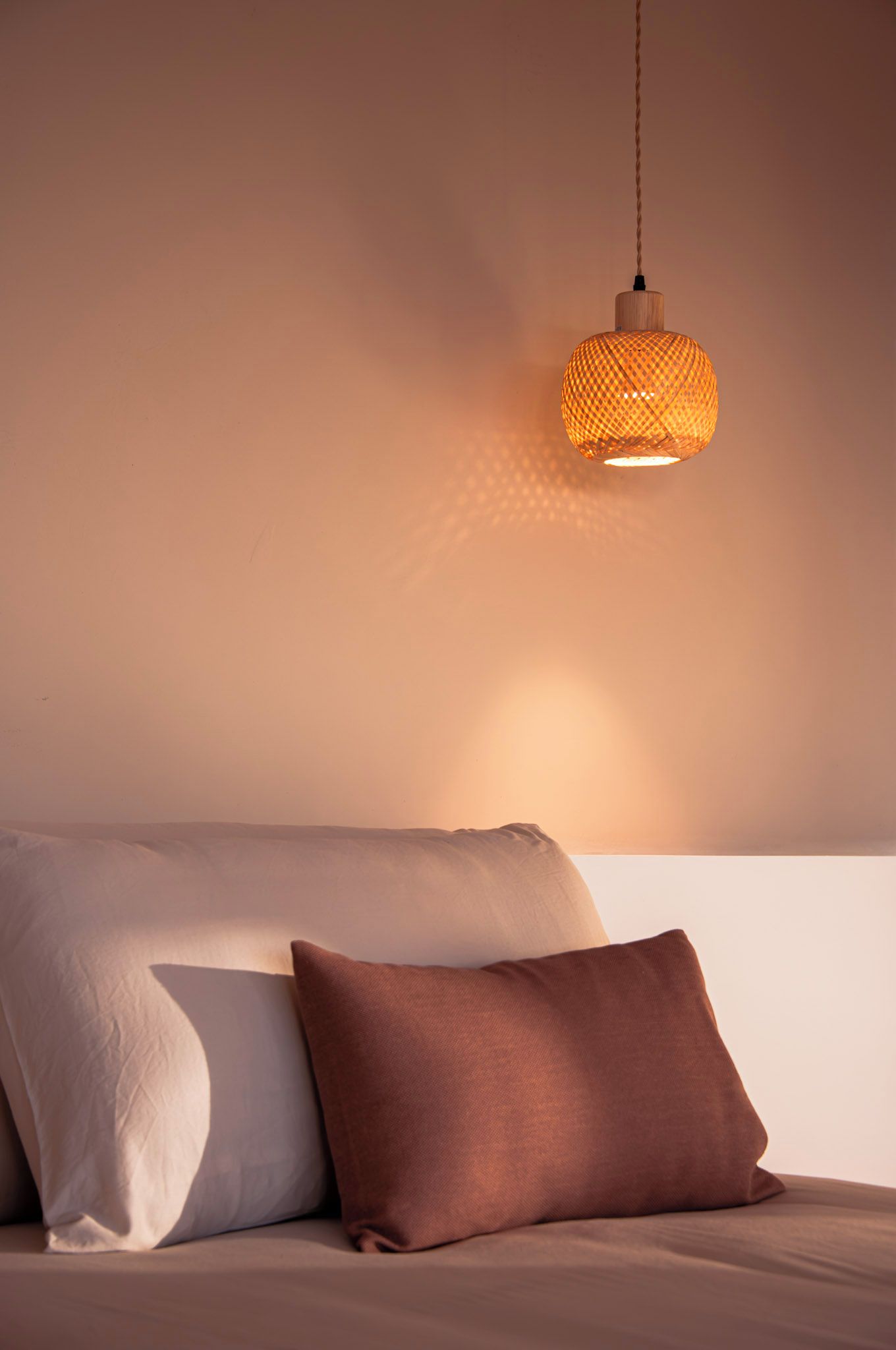 A bed with a single white pillow and a brown pillow, a hanging woven light fixture, and a plain wall.