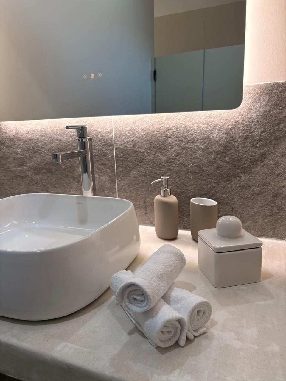 Une salle de bain avec un lavabo rond, un miroir, un porte-serviettes avec des serviettes, un distributeur de savon et un porte-rouleau de papier toilette.