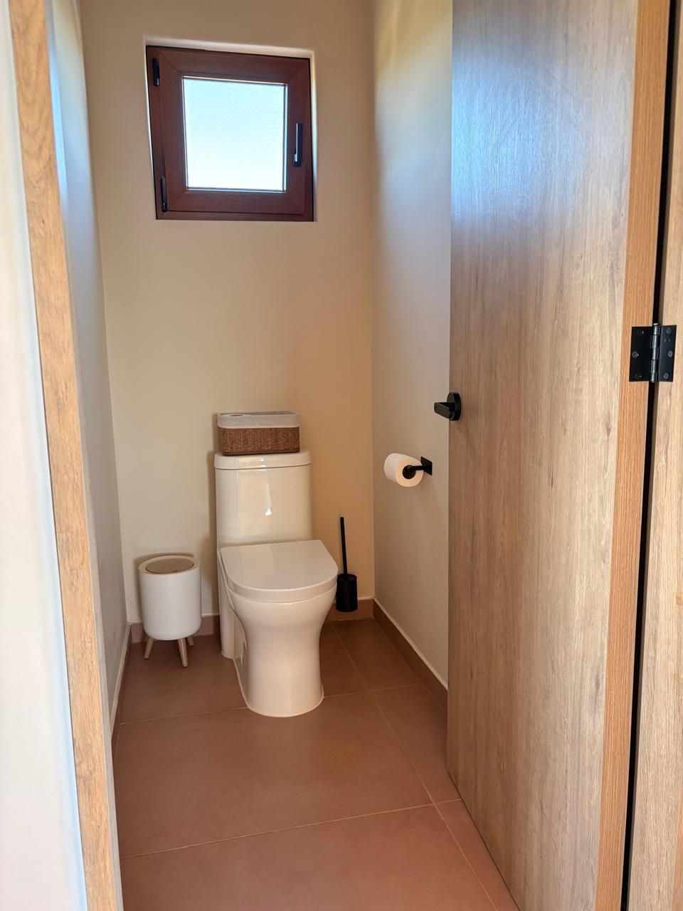 Ein kleines Badezimmer mit Toilette, Toilettenpapier, einem Mülleimer und einem Fenster über der Toilette.