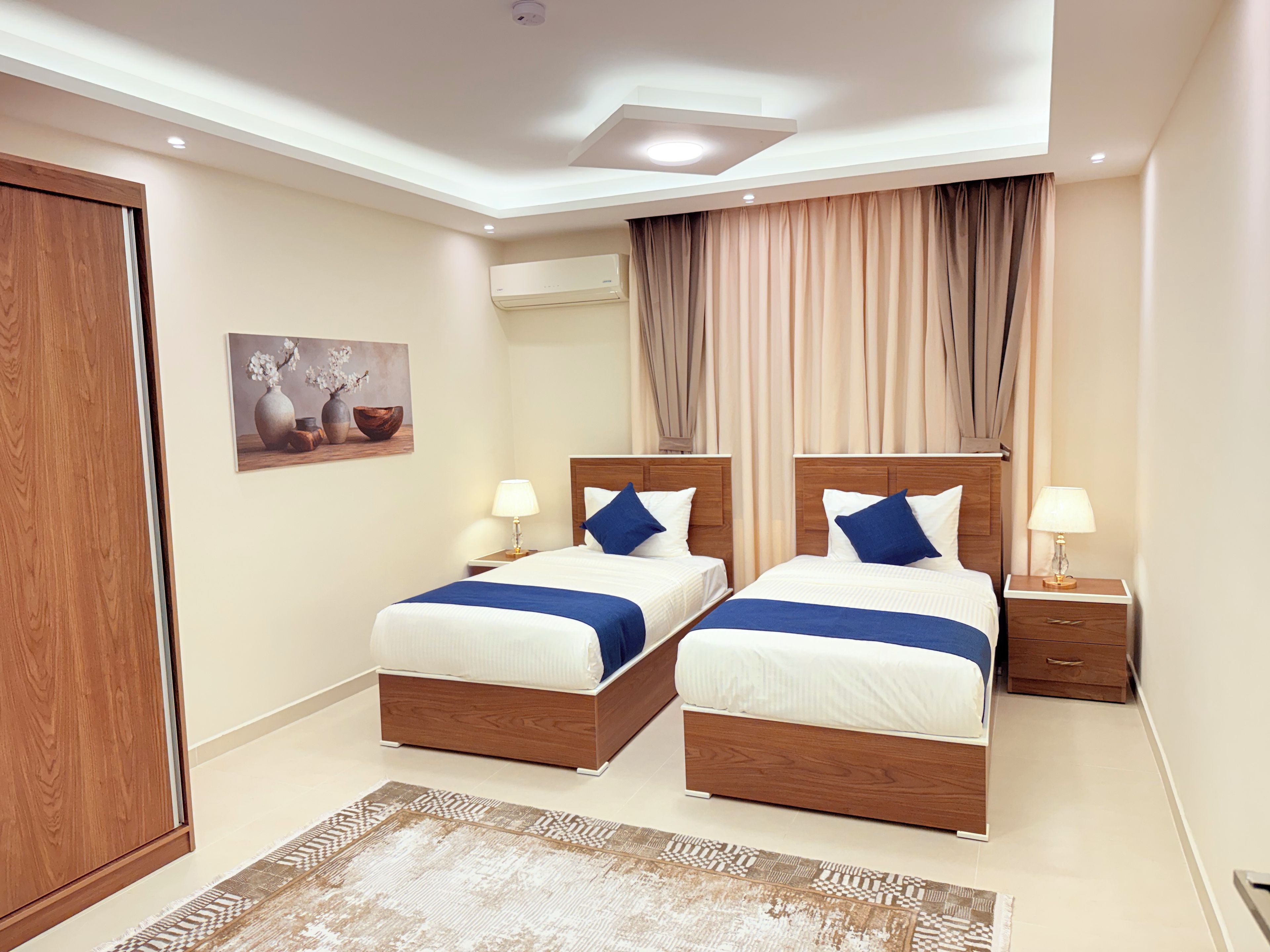 A hotel room with two beds, a wooden wardrobe, a painting, an air conditioning unit, and a window with curtains.