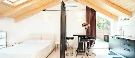 A bedroom with a bed, a desk with two chairs, a window with curtains, and a kitchenette.