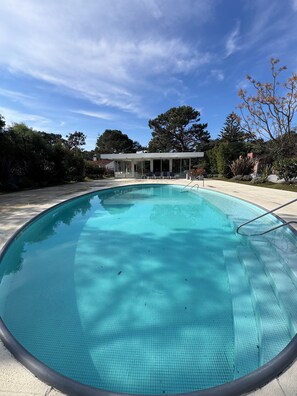 Ein klares Schwimmbecken mit Fliesen umgeben, umgeben von Bäumen und einem modernen Gebäude mit überdachter Terrasse.